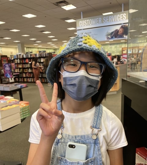 A girl with light brown skin, glasses, and a black bob wearing a bucket hat made of Granny squares and making a peace sign.