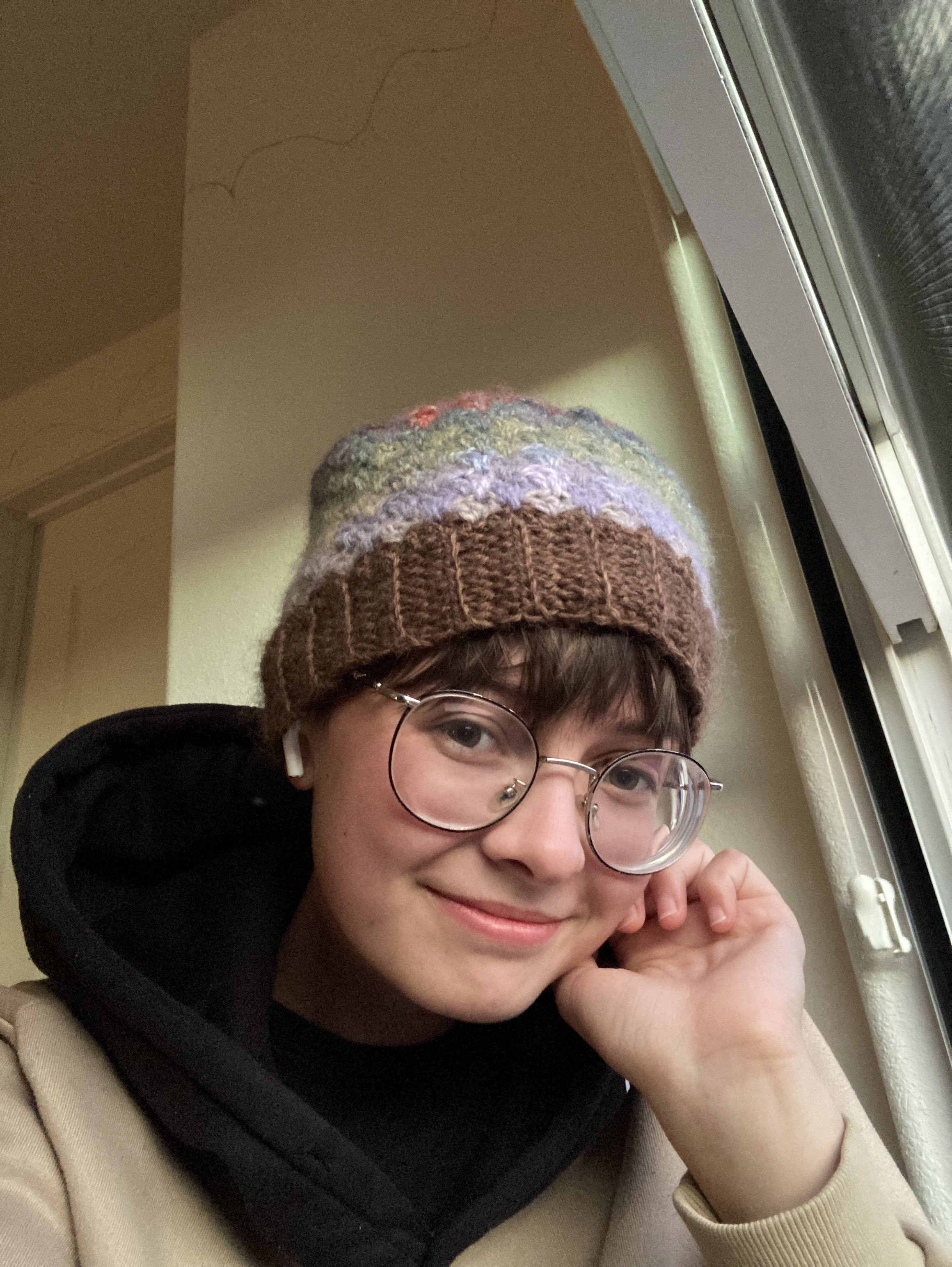 A picture of a young man with light skin, short brown hair, and glasses wearing a crocheted hat in various colors.